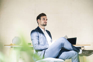 Nachdenkliche junge männliche Fachkraft sitzt mit digitalem Tablet auf einem Stuhl im Büro - MOEF03915