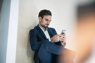 Junger Geschäftsmann, der im Büro sitzend eine Textnachricht über sein Smartphone verschickt - MOEF03916