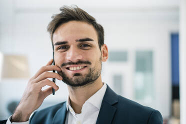 Lächelnder gutaussehender Mann mit Haarstoppeln, der im Büro mit einem Mobiltelefon spricht - MOEF03922