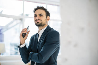 Nachdenklicher junger Geschäftsmann schaut weg, während er sein Smartphone im Büro hält - MOEF03924