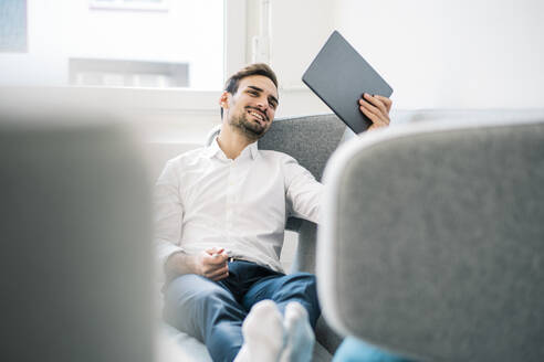 Glücklicher, entspannter männlicher Berufstätiger, der auf dem Sofa im Büro sitzt und ein digitales Tablet liest - MOEF03927