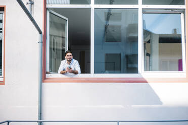 Smiling young businessman holding coffee cup while standing at window - MOEF03930