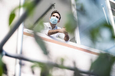 Young businessman wearing protective face mask standing with hands clasped at window - MOEF03934
