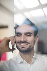 Lächelnder junger Geschäftsmann, der mit einem Smartphone spricht, gesehen durch Glas - MOEF03936