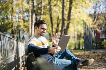 Junger Mann winkt mit der Hand, während er einen Videoanruf über ein digitales Tablet in einem Park an einem sonnigen Tag tätigt - MOEF03939