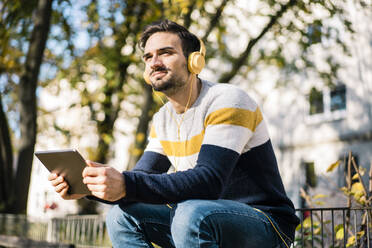 Junger Mann hält digitales Tablet und hört Musik über Kopfhörer im Park an einem sonnigen Tag - MOEF03940