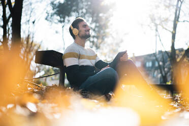 Smiling young man with eyes closed listening music through headphones at park during sunny day - MOEF03945