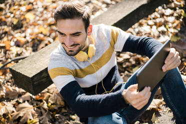 Glücklicher junger Mann mit Kopfhörern und digitalem Tablet, der an einem sonnigen Tag im Park sitzt - MOEF03946