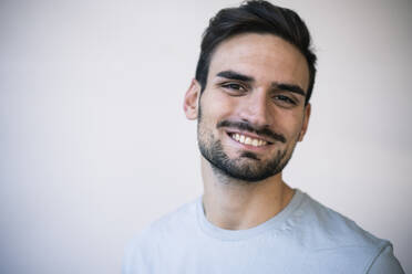 Happy handsome young man with hair stubble in front of wall - MOEF03952