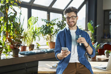 Lächelnder männlicher Berufstätiger, der über sein Mobiltelefon im Büro Textnachrichten verschickt - BSZF01935