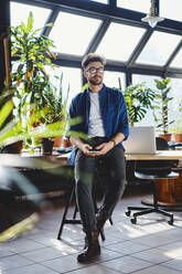 Male professional holding smart phone while sitting on desk in office - BSZF01940