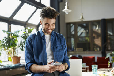 Happy young male professional smiling while using smart phone in office - BSZF01942