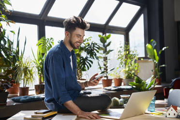 Happy male professional holding smart phone while sitting with laptop at desk in office - BSZF01950