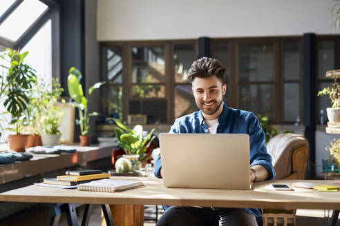 Glücklicher Geschäftsmann arbeitet am Laptop am Schreibtisch im Büro - BSZF01955