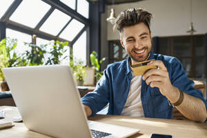 Junger Geschäftsmann, der mit seiner Kreditkarte bezahlt, während er einen Laptop im Büro benutzt - BSZF01959