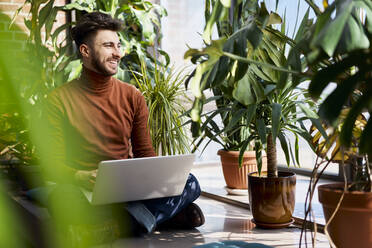 Happy young man sitting with laptop on window sill at home - BSZF01976