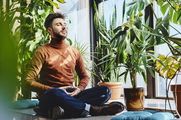 Young man meditating by plants on window sill at home - BSZF01977