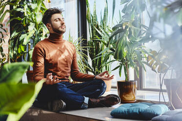 Man sitting cross-legged while meditating on window sill at home - BSZF01978