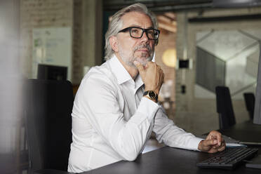 Thoughtful businessman with hand on chin sitting at desk in office - RBF08325