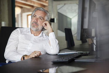 Thoughtful male professional with gray hair sitting at desk in office - RBF08326