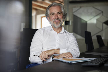 Smiling bearded male professional with documents sitting at desk in office - RBF08329