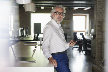 Mature businessman holding smart phone leaning on desk at office - RBF08334