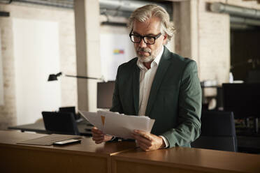 Geschäftsmann mit grauem Haar analysiert Dokumente, während er an einem Tisch im Büro steht - RBF08343