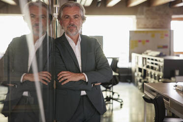 Mature businessman standing with arms crossed by glass in office - RBF08347