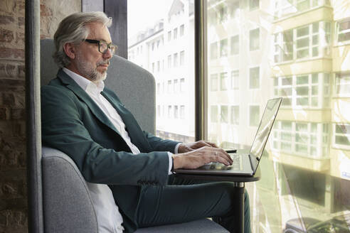 Reife männliche professionelle Arbeit mit Laptop, während auf dem Sofa durch Glasfenster im Büro sitzen - RBF08349