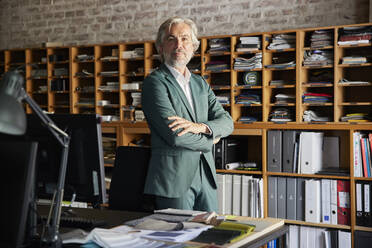 Confident male professional standing with arms crossed by desk in office - RBF08355