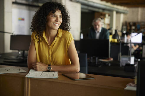Smiling curly haired businesswoman with documents and digital tablet leaning on table at office - RBF08362