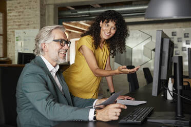Lächelnde Geschäftsfrau gestikuliert, während sie mit einem männlichen Kollegen im Büro über den Computer diskutiert - RBF08364