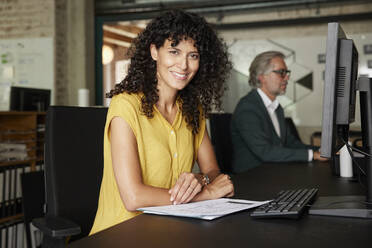 Smiling female professional with documents sitting at desk in office - RBF08365