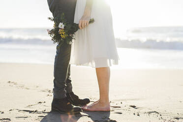 Braut hält Blumenstrauß, während sie mit dem Bräutigam am Strand steht - AFVF09153