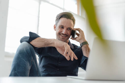 Lächelnde männliche Fachkraft, die am Arbeitsplatz mit einem Smartphone spricht - JOSEF05488