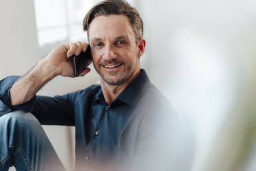 Lächelnde reife männliche Fachkraft, die im Büro mit einem Mobiltelefon spricht - JOSEF05500