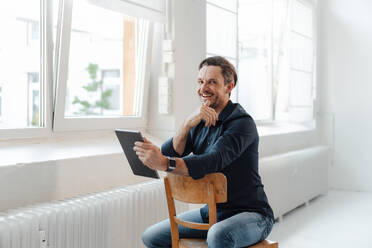 Lächelnder Geschäftsmann, der ein digitales Tablet hält und auf einem Stuhl im Büro sitzt - JOSEF05501