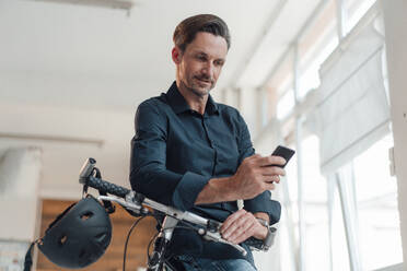 Älterer männlicher Berufstätiger, der sein Smartphone auf dem Fahrrad am Arbeitsplatz benutzt - JOSEF05515