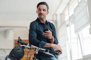 Mature man with smart phone and bicycle in office - JOSEF05516