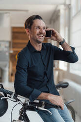 Happy mature businessman talking on smart phone while sitting on bicycle in office - JOSEF05517