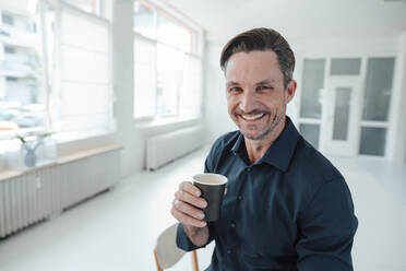 Lächelnder Geschäftsmann mit Kaffee im Einwegbecher am Arbeitsplatz - JOSEF05534