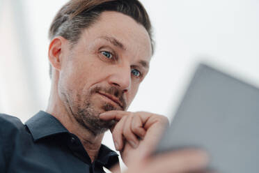 Businessman with hand on chin looking at digital tablet in office - JOSEF05542