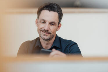 Lächelnder männlicher Berufstätiger, der eine Textnachricht auf einem Smartphone im Büro schreibt - JOSEF05551