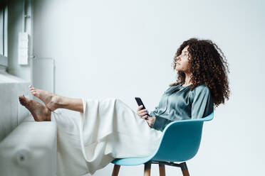 Nachdenkliche Geschäftsfrau mit Smartphone auf einem Stuhl im Büro sitzend - JOSEF05573