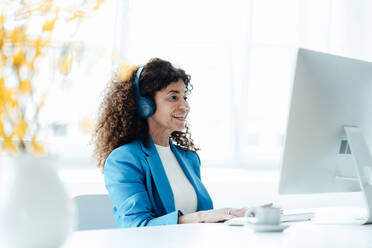 Mature businesswoman wearing headphones while working at desk in office - JOSEF05597