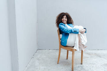Geschäftsfrau, die ihre Knie umarmt und auf einem Stuhl vor einer grauen Wand sitzt - JOSEF05604