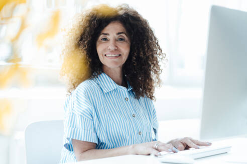 Mature businesswoman with curly hair sitting at desk in office - JOSEF05617