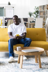 Smiling man with smart phone sitting on sofa at home - GIOF13335