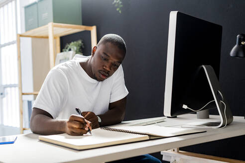 Male freelancer writing in spiral notebook at desk in studio - GIOF13415