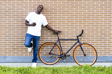Mann mit Smartphone und Fahrrad vor einer Backsteinmauer stehend - GIOF13432